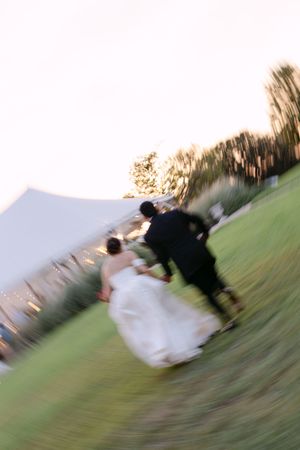 Blurred motion photo of bride and groom running across green lawn toward white tent at outdoor wedding reception.