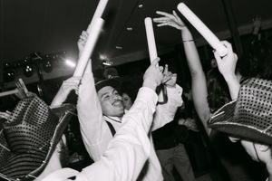 Man in white suit celebrates with glow sticks at a lively party in black and white photo.