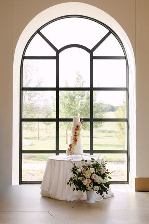 Elegant wedding cake on draped table with floral arrangement before arched window overlooking lush garden.
