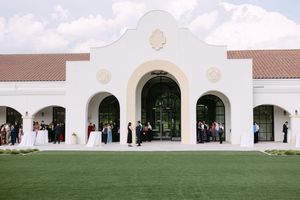 Elegant white Spanish-style wedding venue with arched entryways and guests gathered on lush green lawn.