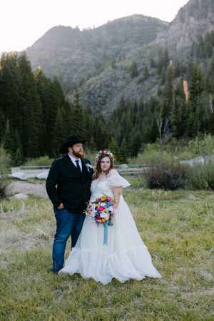 Bride and groom pose in mountain meadow, she wears floral crown and white gown, he wears black cowboy hat and suit.