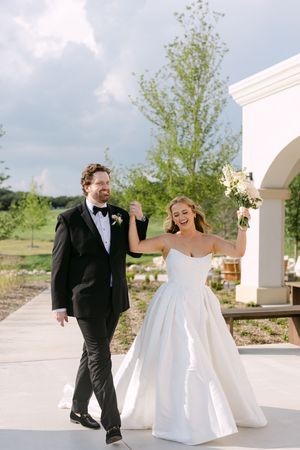 Joyful bride and groom walking together outdoors, bride in strapless ballgown holding bouquet aloft.
