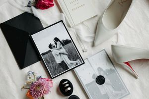 Black and white wedding flat lay with couple photo, white heels, floral bouquet, rings, and elegant stationery on white background.