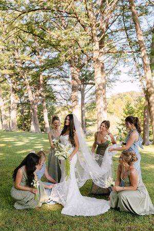 Bride in white gown and veil surrounded by bridesmaids in sage green dresses on lush green lawn with tall trees.