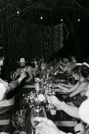 Black and white photo of guests toasting at an outdoor evening wedding reception with string lights overhead.