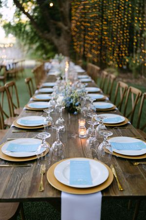 Elegant outdoor wedding reception with long wooden table, gold chargers, blue menus, crystal glasses, and fairy light backdrop.