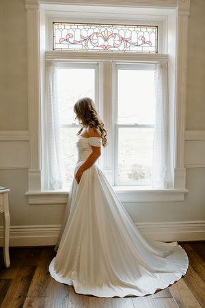 A bride in a flowing white ballgown stands elegantly near large windows in a bright room with hardwood floors.