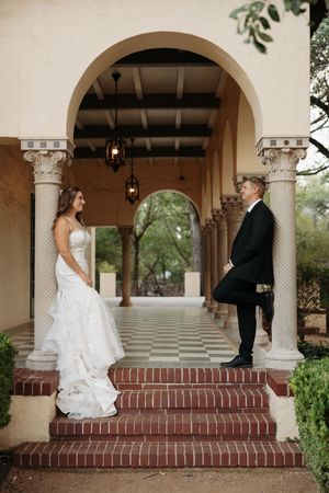 A bride and groom share a romantic moment on brick steps under an elegant arched colonnade.