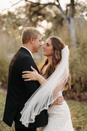 A romantic wedding couple embraces outdoors during golden hour with the bride wearing a flowing veil and lace dress.
