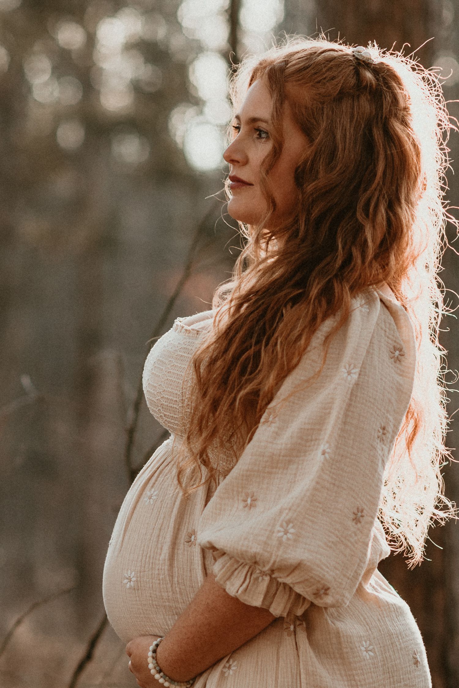 Sunlit portrait of a woman in a white dress holding her growing baby bump.