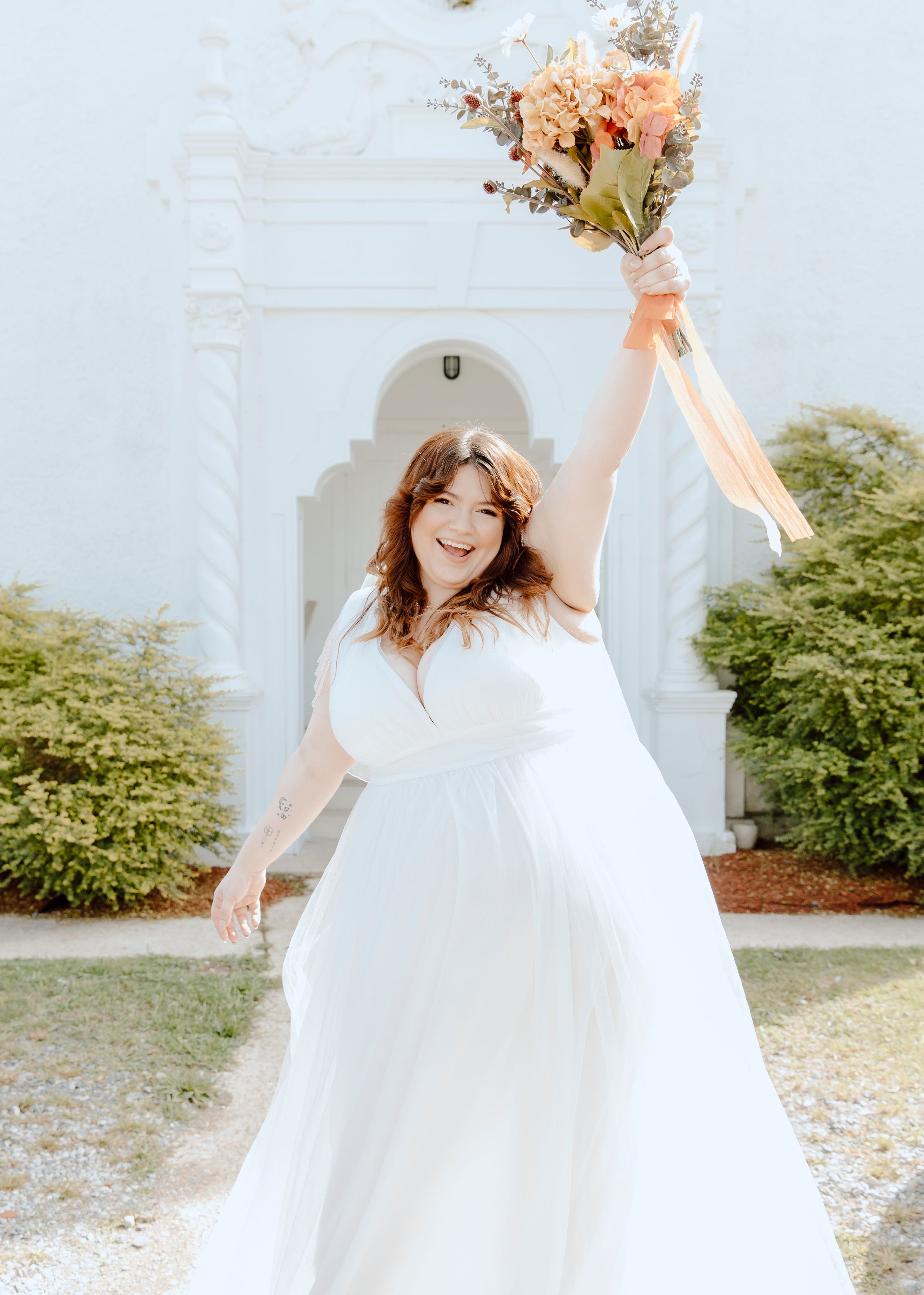 A bride in a white wedding dress celebrates joyfully with her bouquet outdoors.