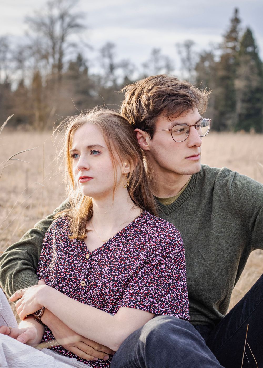 man and woman sit in field, she leans against his chest, his arm is around her and he look right while she looks left