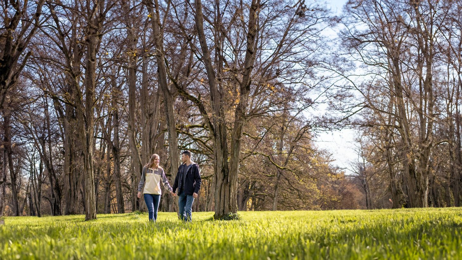 man holds hand of woman and leads her toward camera as they walk in a large field with few towering trees