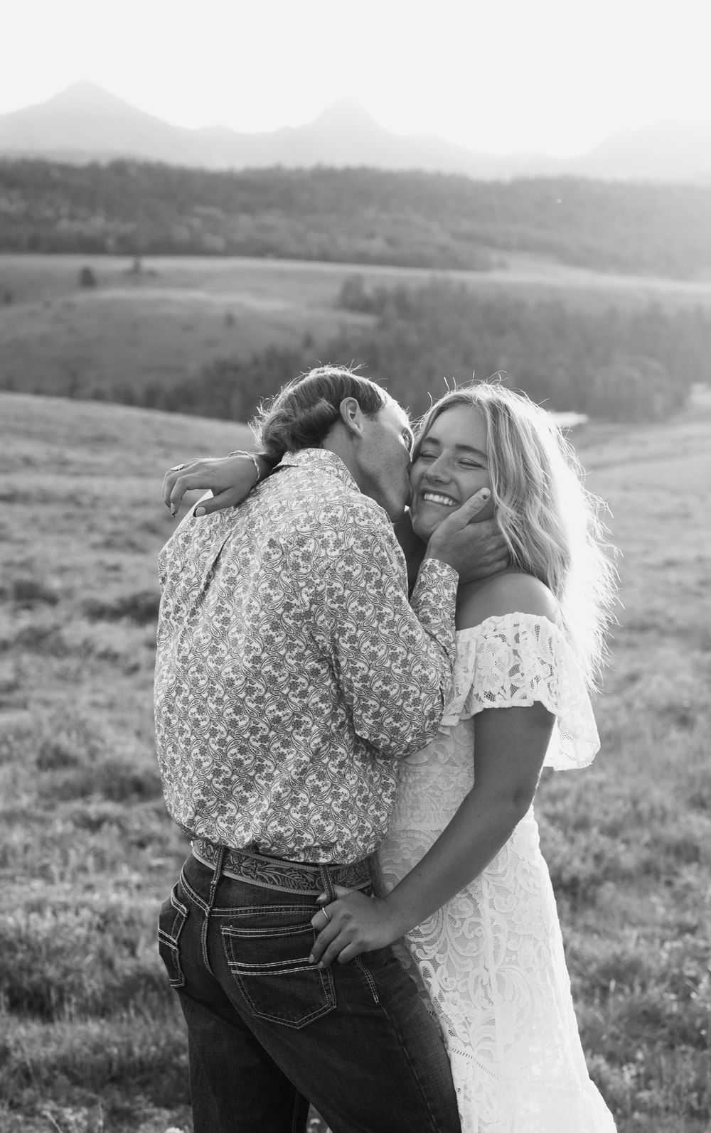 A romantic couple embraces in a scenic mountain meadow setting captured in black and white photography.