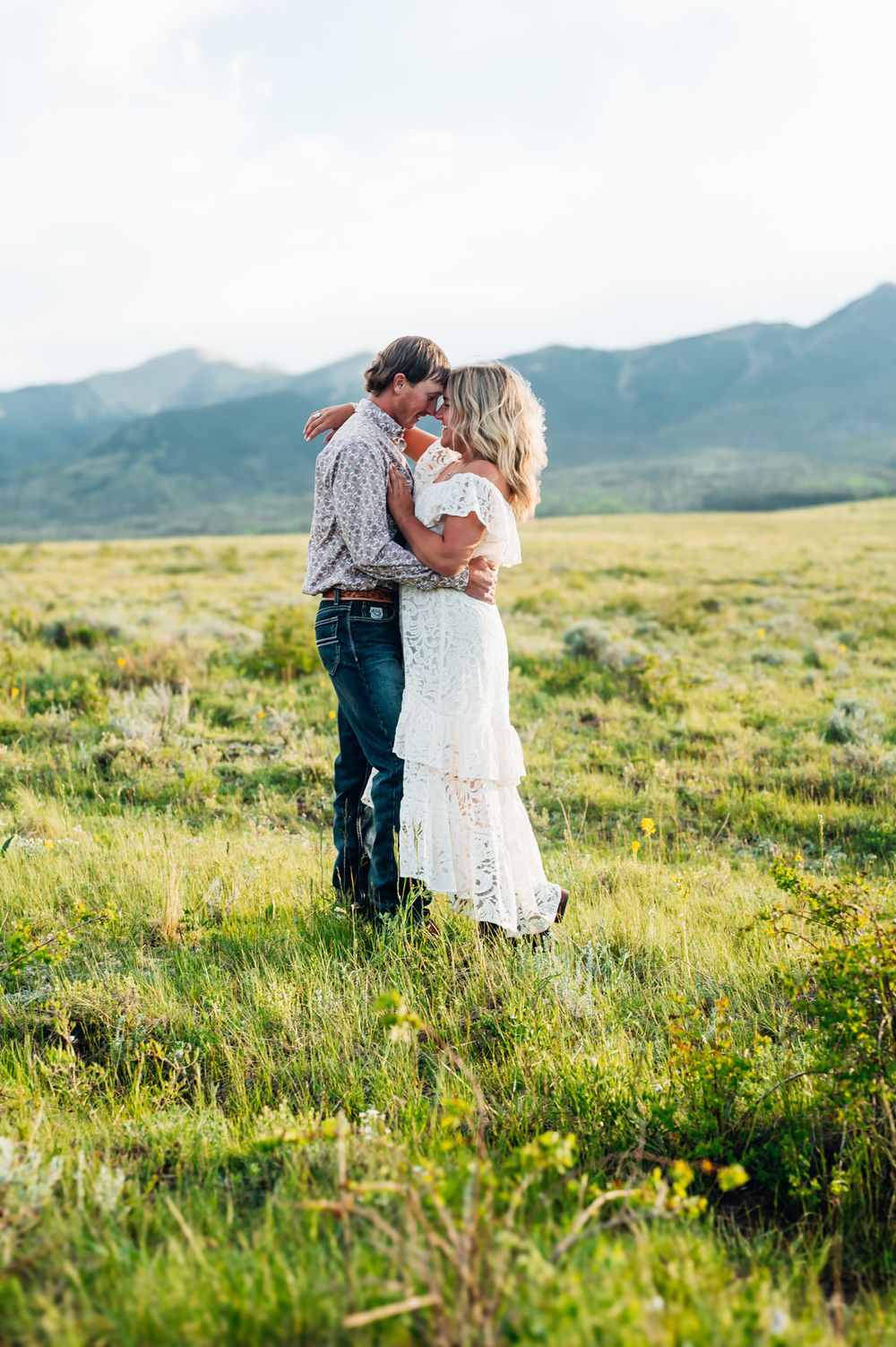 A romantic couple embraces in a lush mountain meadow during golden hour with mountains in the background.