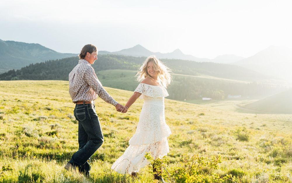 A couple holds hands while dancing together in a sunlit mountain meadow with wildflowers.