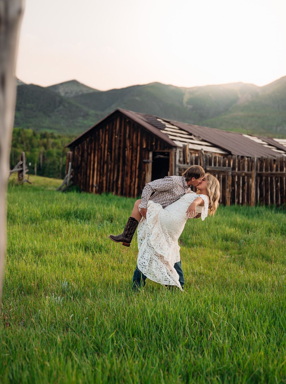 A couple dances playfully in a green meadow with rustic wooden barns in the background during sunset.