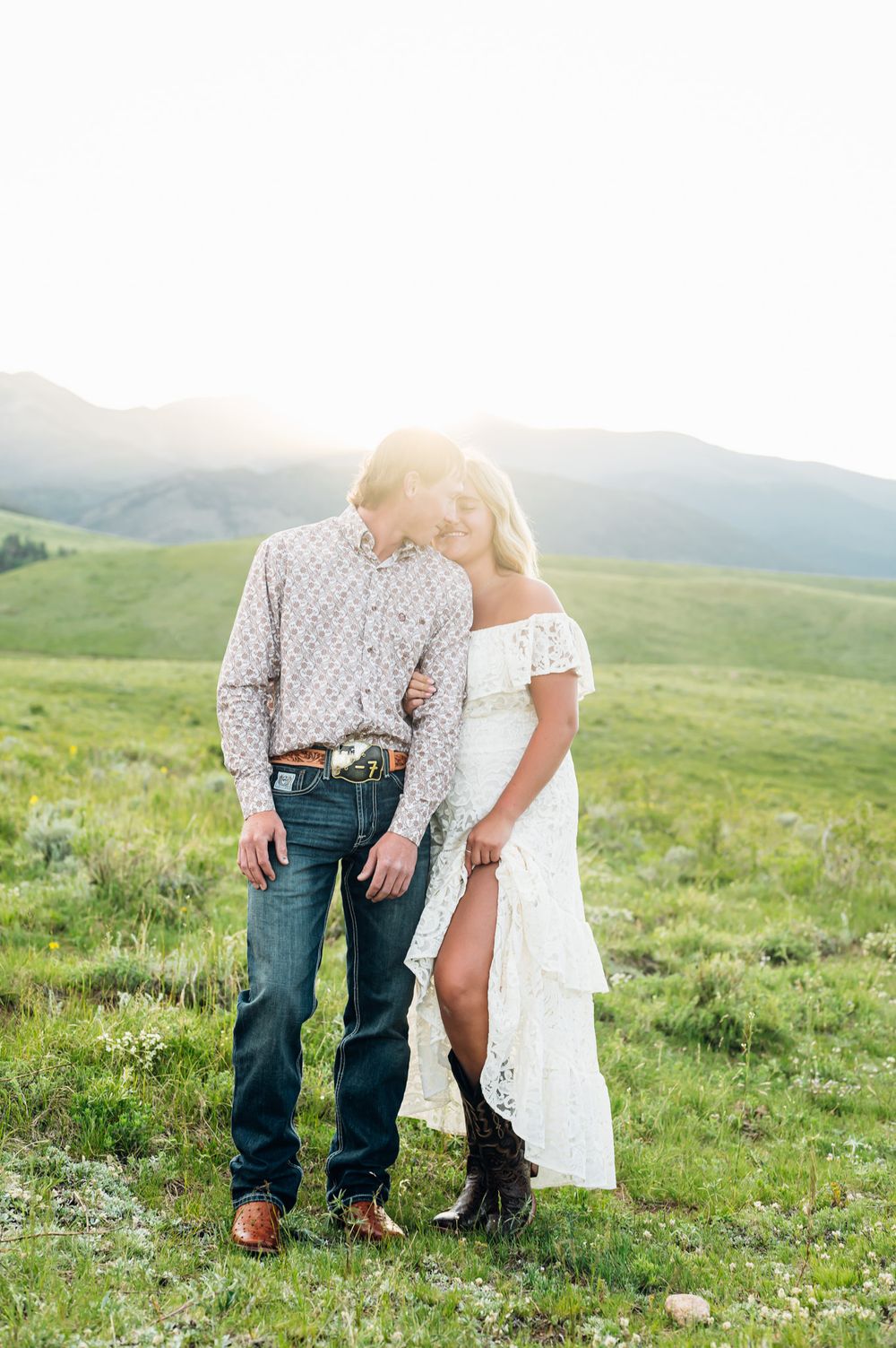 Couple shares intimate moment on mountainside meadow with warm sunlight filtering through rolling hills.