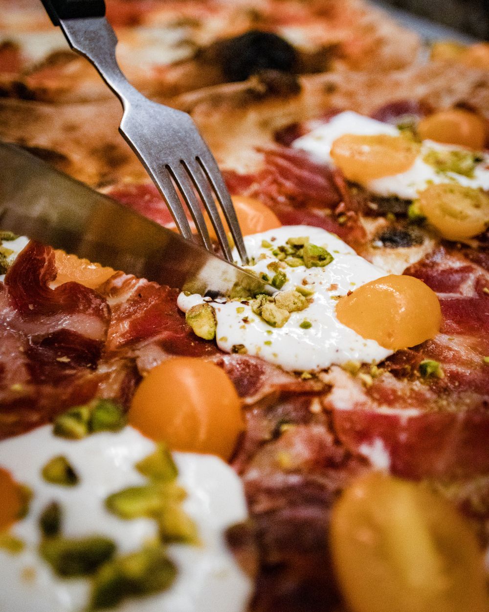 Close-up of pizza with prosciutto, burrata cheese, pistachios and cherry tomatoes being cut with utensils.