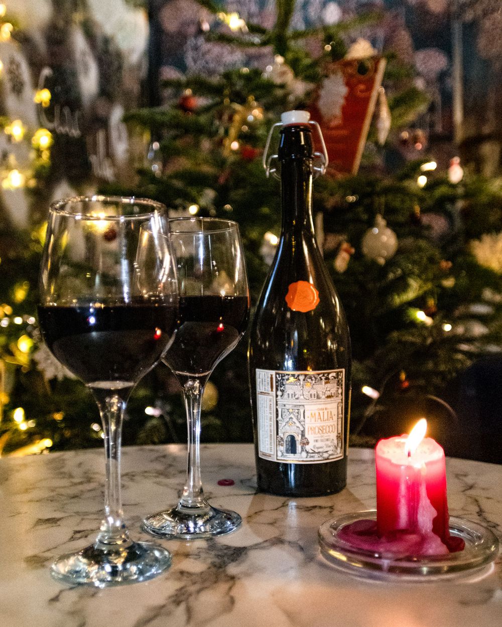Wine bottle and glasses with red candle against blurred Christmas tree lights.