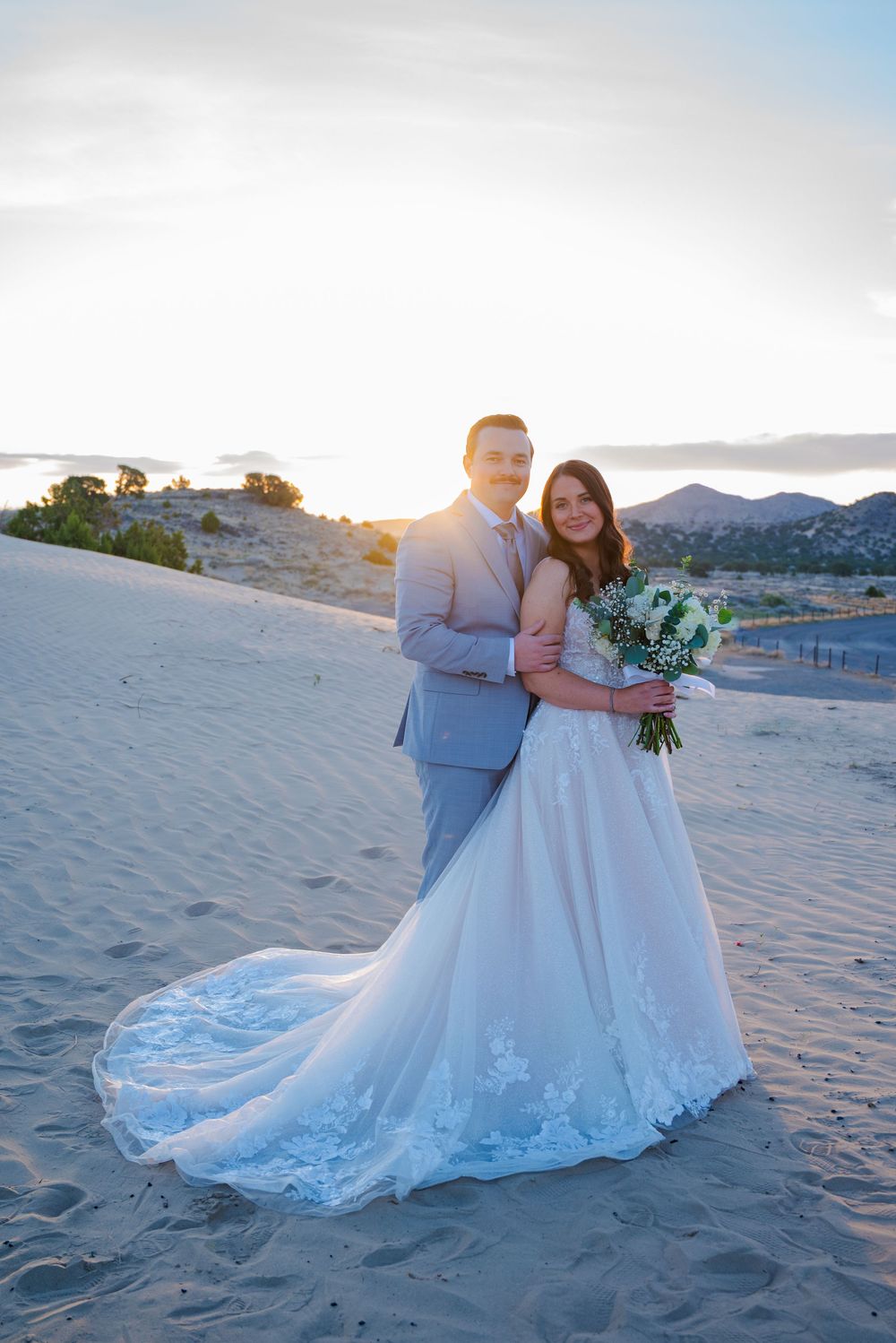 Bride holding floral bouquet poses with groom on sandy dunes in soft golden sunset light.