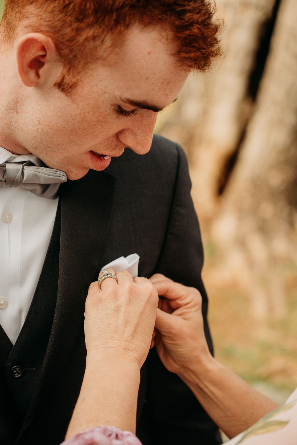 Red-haired groom in black tuxedo and gray bow tie has pocket square adjusted by a loved one.
