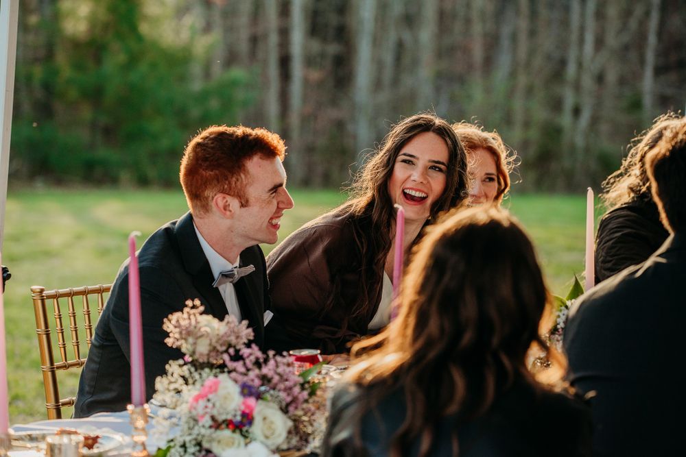Bride and groom laughing joyfully with guests at decorated outdoor wedding reception dinner table.