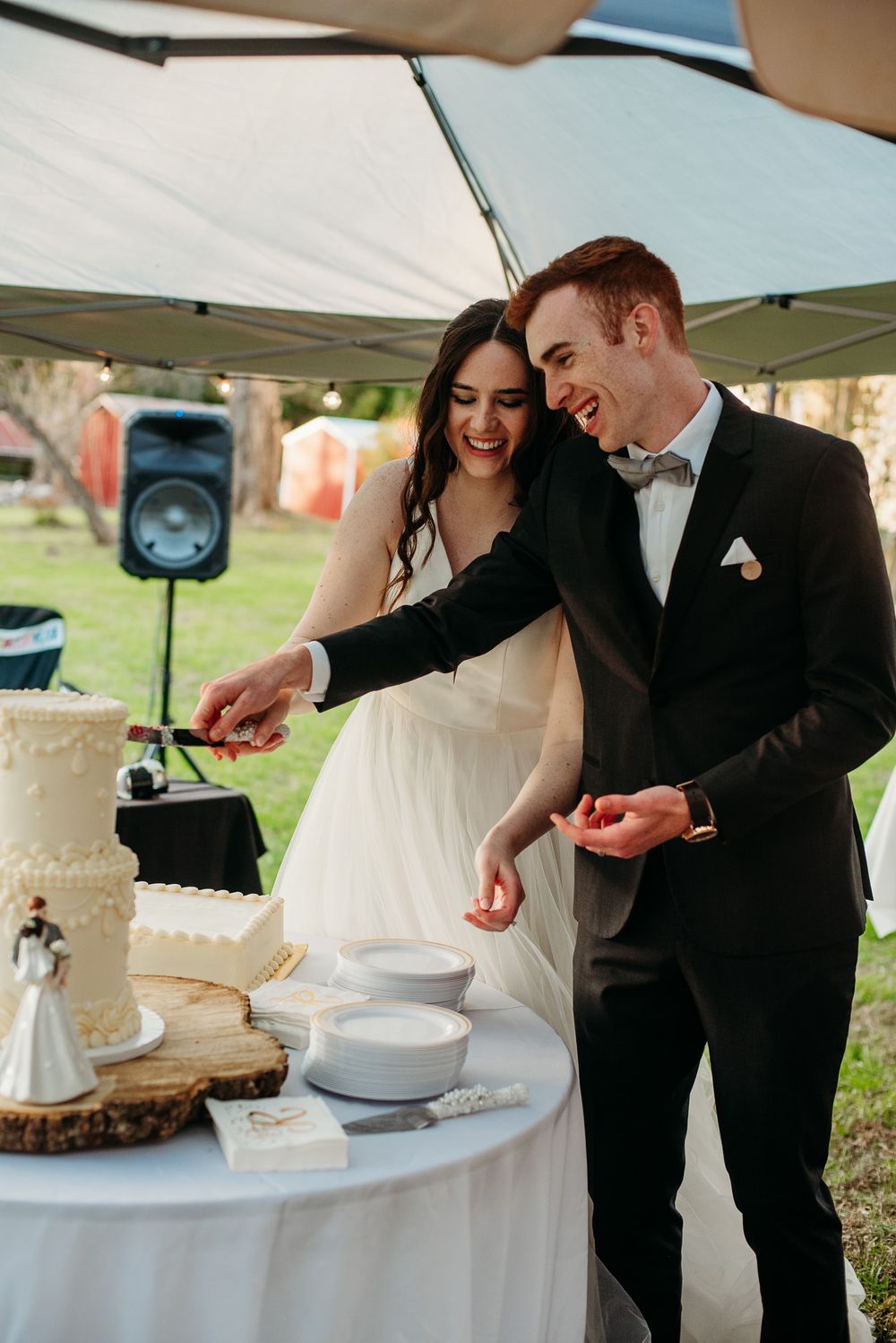 Laughing bride and groom cutting their white wedding cake together under an outdoor reception tent.