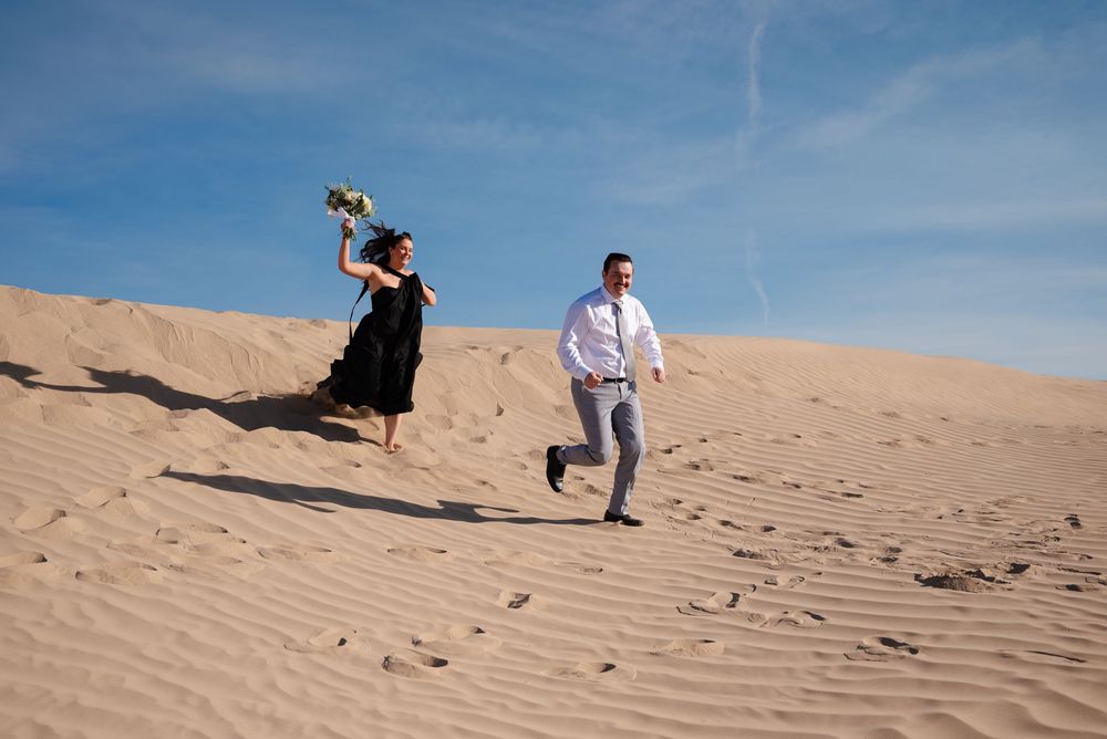 Bride in black dress playfully chasing groom on vast sandy desert dunes under blue sky.