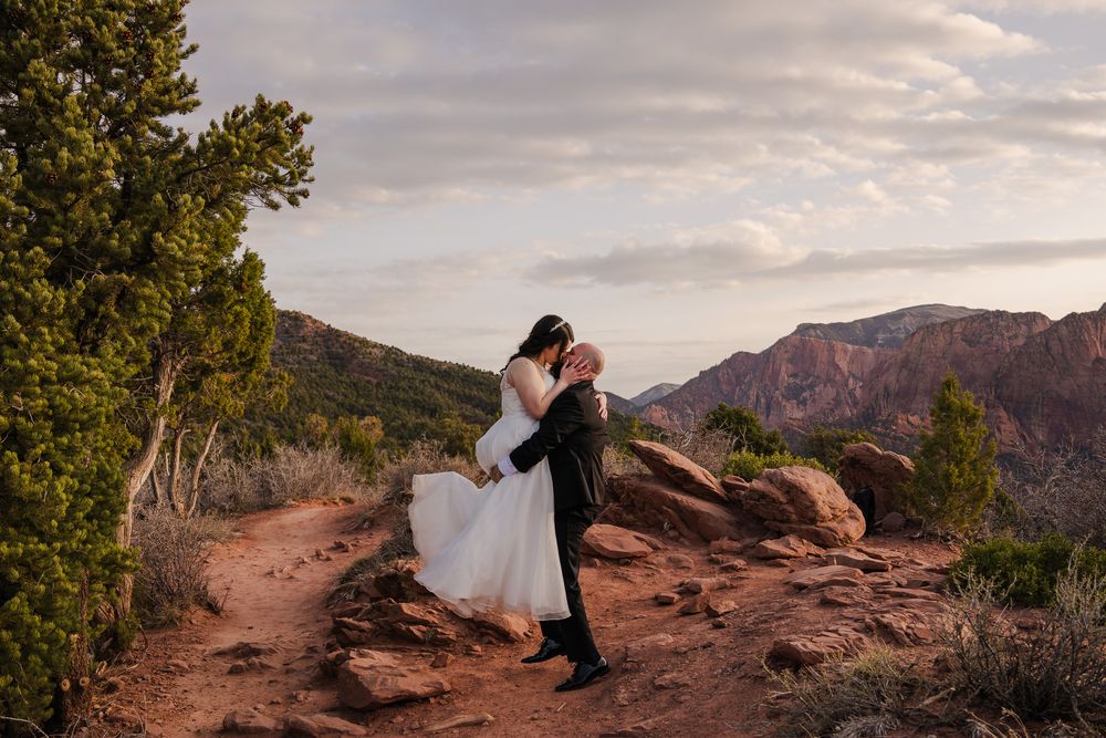 Groom lifting bride in white dress on scenic red dirt trail with canyon mountain views.