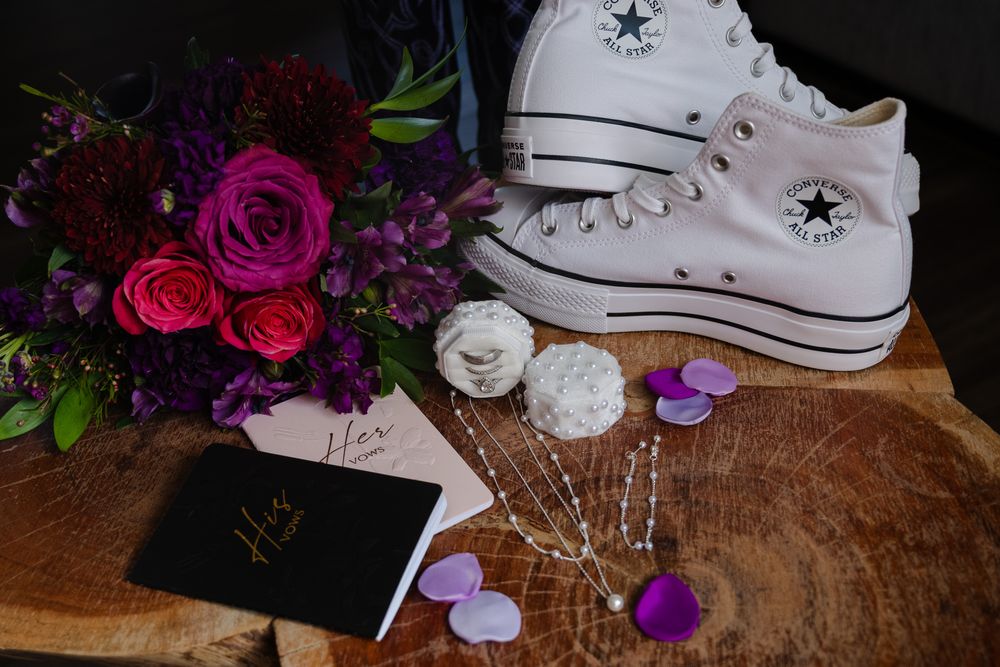Wedding flat lay featuring white Converse sneakers, purple bouquet, vow books, and decorative hair pin.