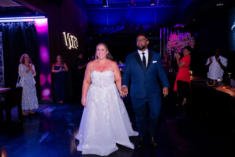 Interracial bride and groom making a grand entrance at their wedding reception in a dark, neon-lit venue.