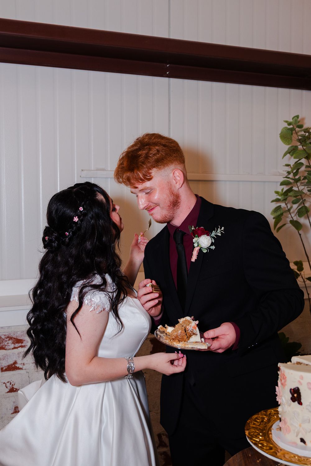 Bride feeds cake to smiling red-haired groom beside white wedding cake at reception.