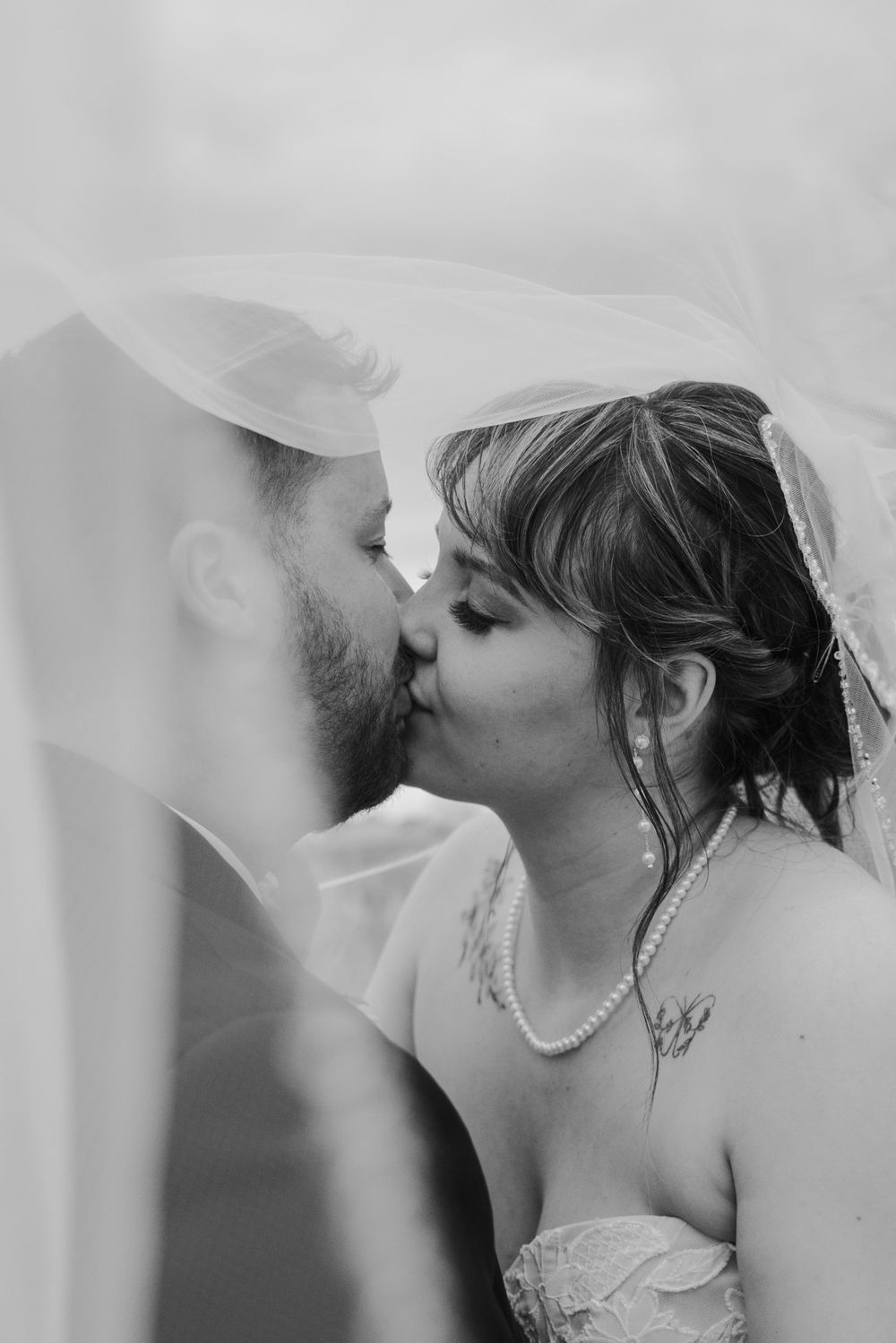 Intimate black and white photo of bride and groom kissing beneath flowing wedding veil outdoors.