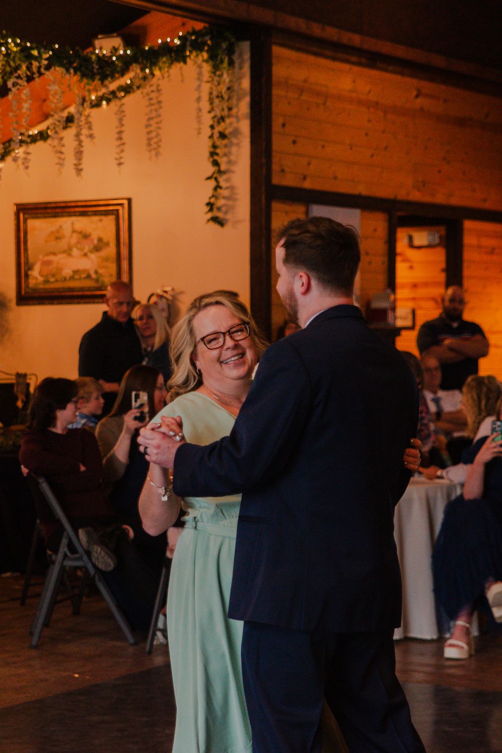 Mother of groom laughing happily while dancing with groom at warm-lit barn wedding reception.