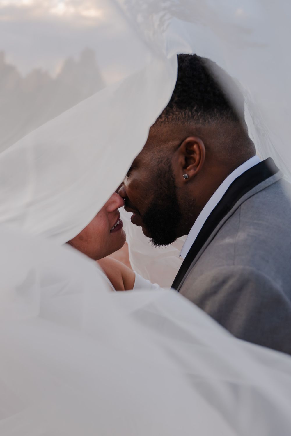 Intimate close-up of interracial bride and groom nearly kissing beneath flowing white veil.