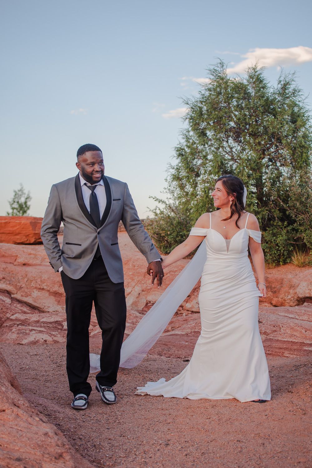 Bride and groom walking hand in hand across red rock terrain at sunset in a desert landscape.