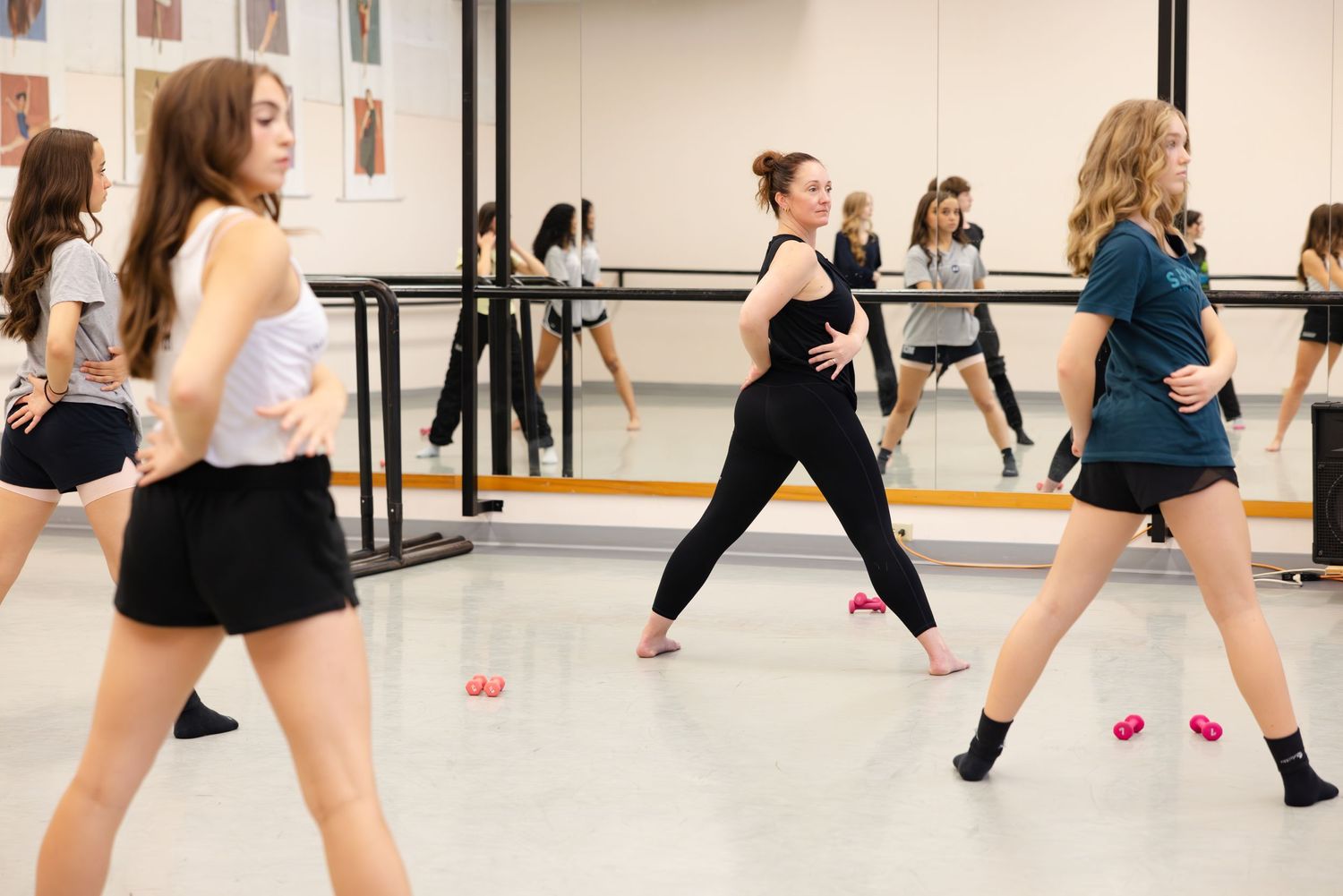 Dance students practice choreography in a studio with mirrors and ballet barres while wearing athletic attire during class.