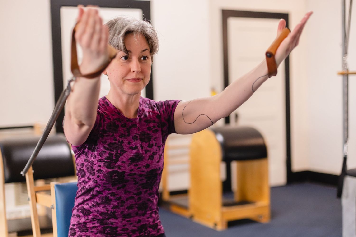 Someone in a purple and black patterned top poses with raised arms during a Pilates or fitness session at a studio.