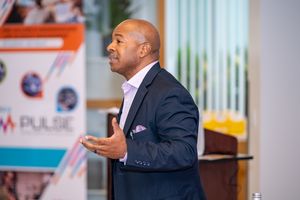 Professional speaker in dark suit gesturing while giving presentation at corporate event.