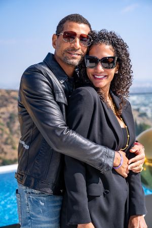A couple in black outfits embraces while posing near an infinity pool with scenic mountain views.