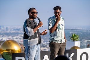 Two people speaking into microphones at an outdoor event with cityscape backdrop.