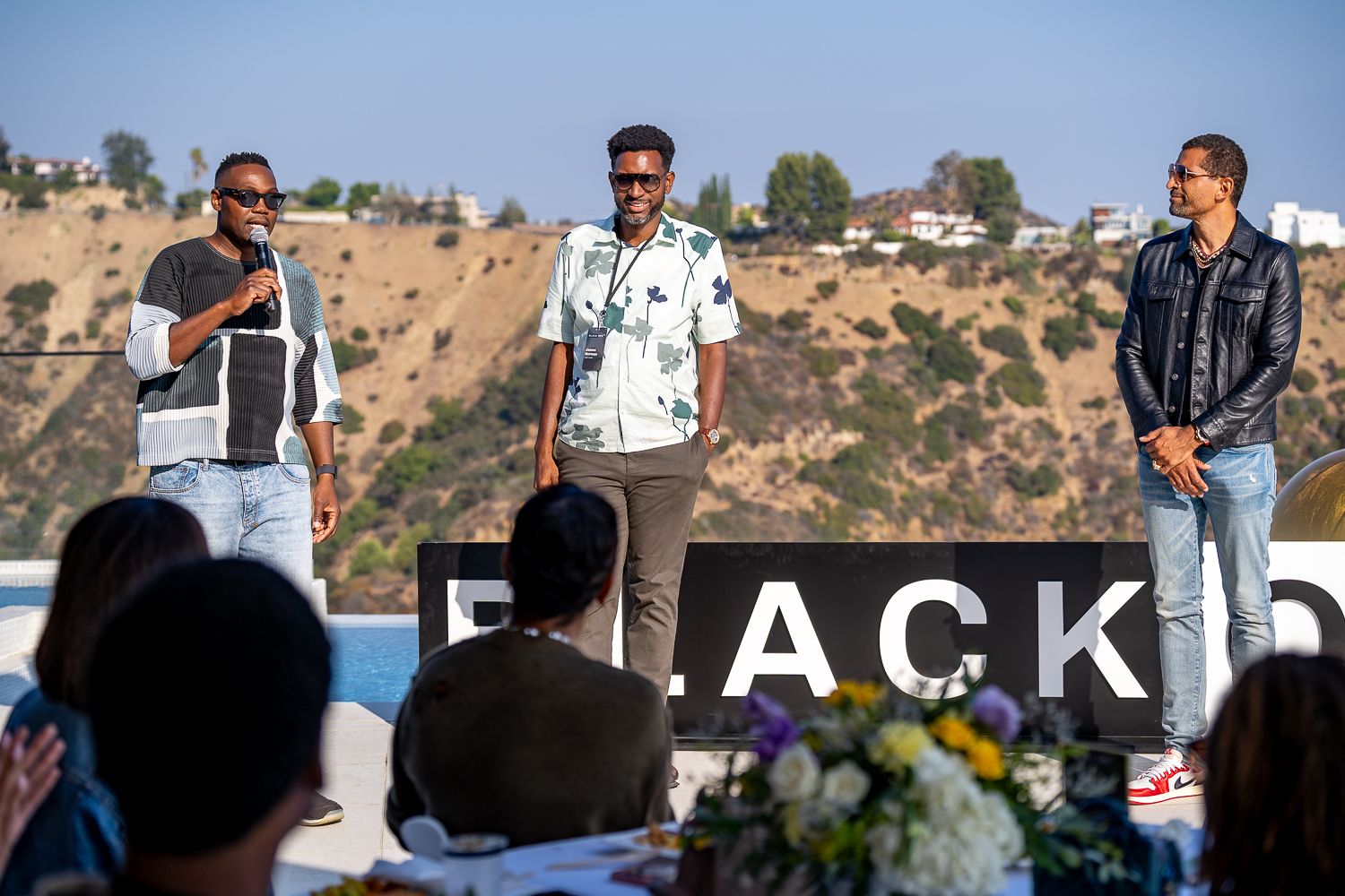 Three speakers present on an outdoor stage with hills in the background.