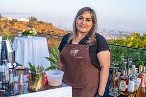 A smiling bartender in brown apron stands behind an outdoor bar setup with cocktail supplies.