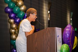 Speaker at podium with decorative balloon arch display in purple and gold.