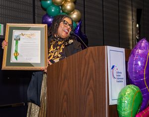 Speaker at podium displaying award certificate surrounded by colorful balloons at professional event.
