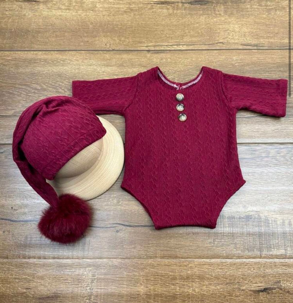 A burgundy knitted baby sweater with pom pom details laid out on a wooden surface.