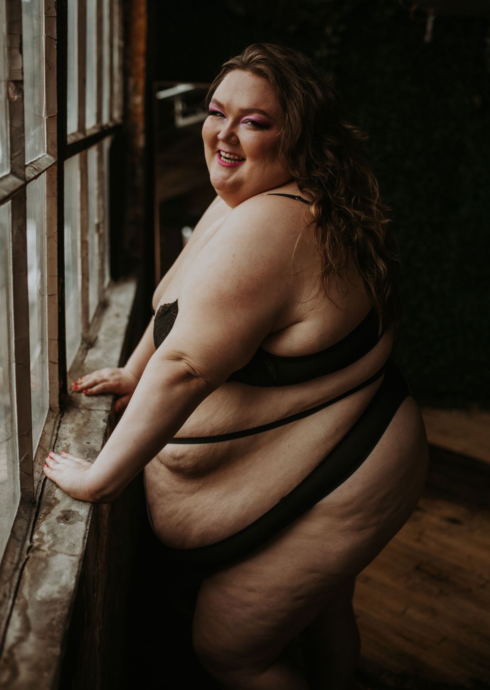 Plus-size woman in black lingerie posing confidently by a rustic window, smiling over her shoulder.
