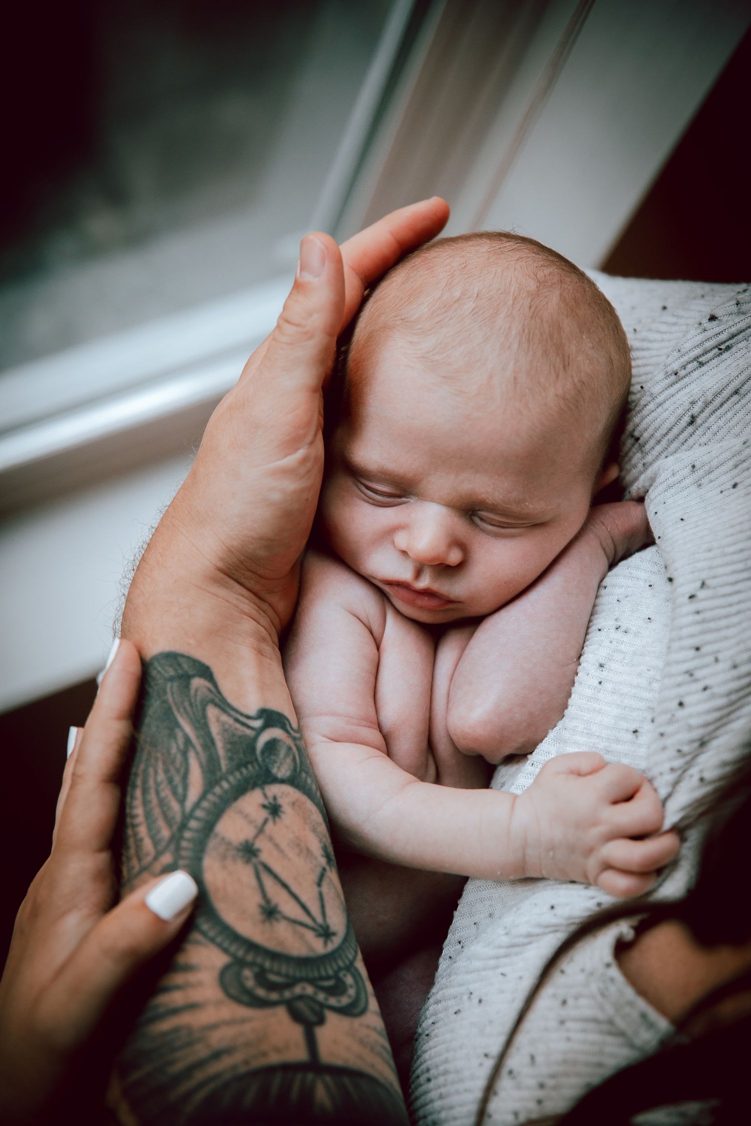 Sleeping newborn resting peacefully on tattooed hands near a window.