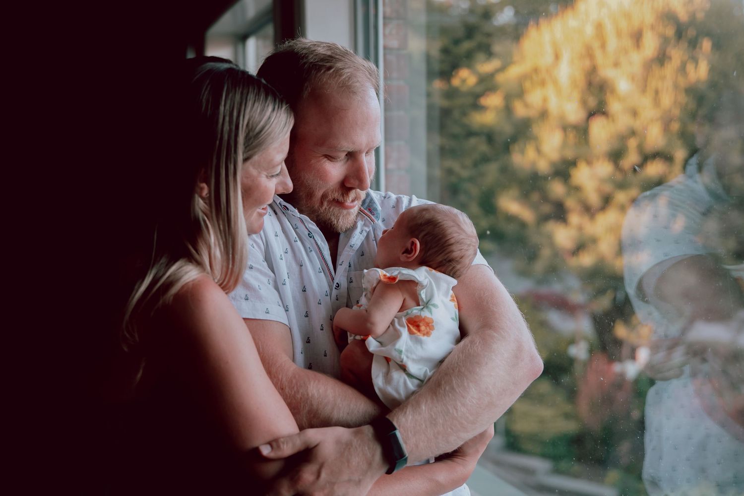 New parents gazing lovingly at their newborn baby by a sunlit window.